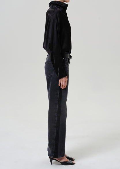 Woman wearing dark grey wide leg jeans with a black draped neck top viewed from the side on a grey background.