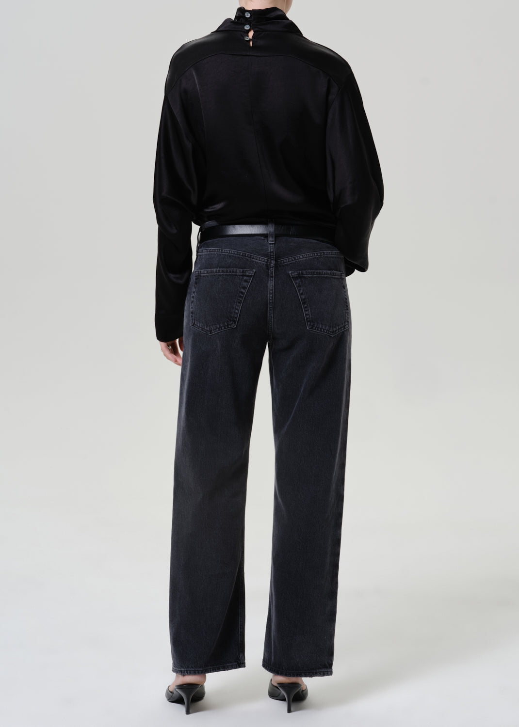 Woman wearing dark grey wide leg jeans with a black draped neck top viewed from the back on a grey background.