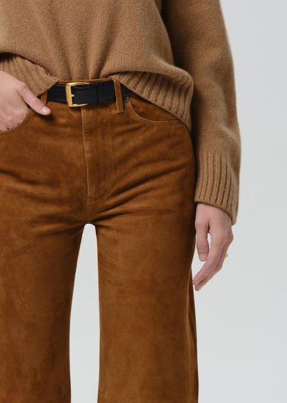 A close-up of a person wearing brown suede trousers, a tan ribbed knit sweater, and a black belt with a gold buckle, against a grey background.