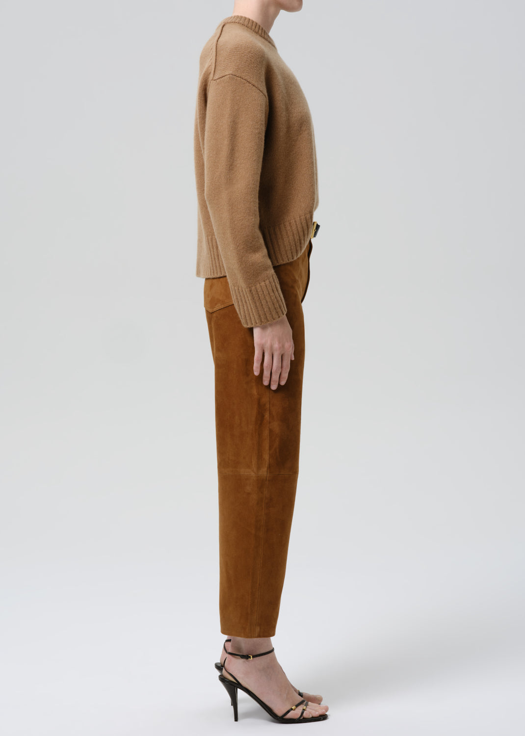 A model shown from the side wearing a camel-colored knit sweater and matching brown suede trousers, paired with black strappy high-heeled sandals, against a grey background.