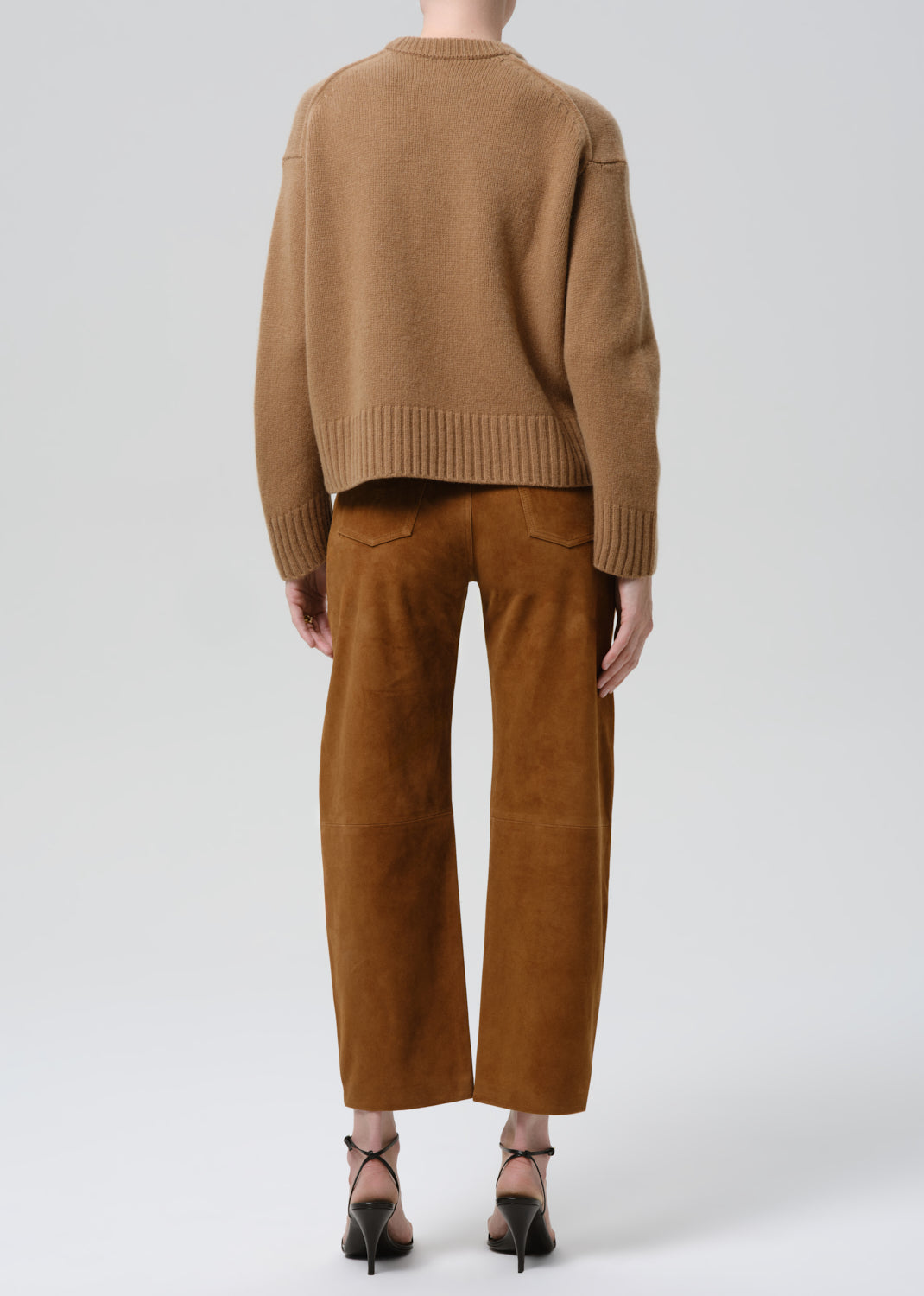 A model shown from the back wearing a cropped tan knit sweater and matching wide-leg brown suede trousers, paired with black high-heeled sandals, against a grey background.