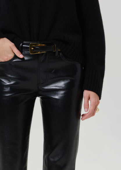 Close-up of a person wearing shiny black faux-leather pants with a black belt featuring a gold buckle, paired with a black ribbed sweater against a light gray studio background.
