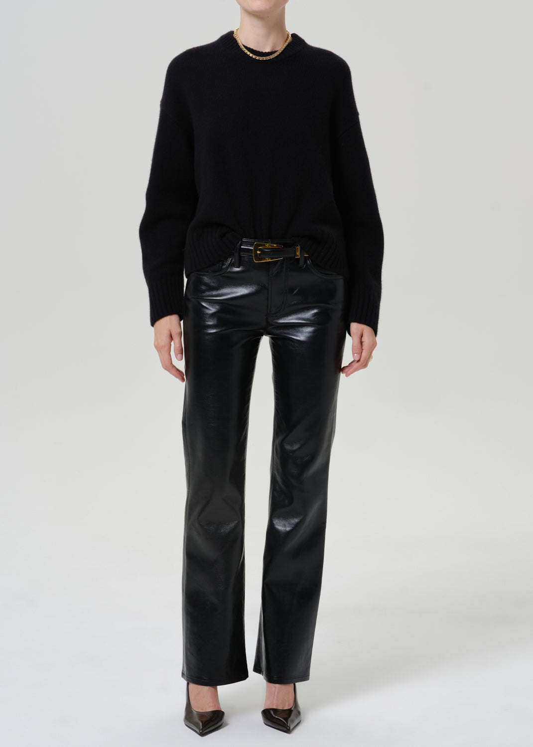 A model wearing a black crewneck sweater, black leather pants with a gold-tone belt, and black pointed-toe heels, photographed against a seamless light gray studio background.