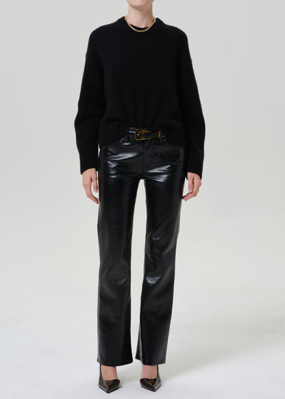 A model wearing a black crewneck sweater, black leather pants with a gold-tone belt, and black pointed-toe heels, photographed against a seamless light gray studio background.