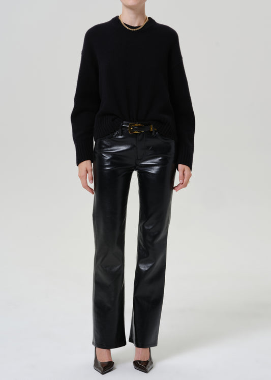 A model wearing a black crewneck sweater, black leather pants with a gold-tone belt, and black pointed-toe heels, photographed against a seamless light gray studio background.