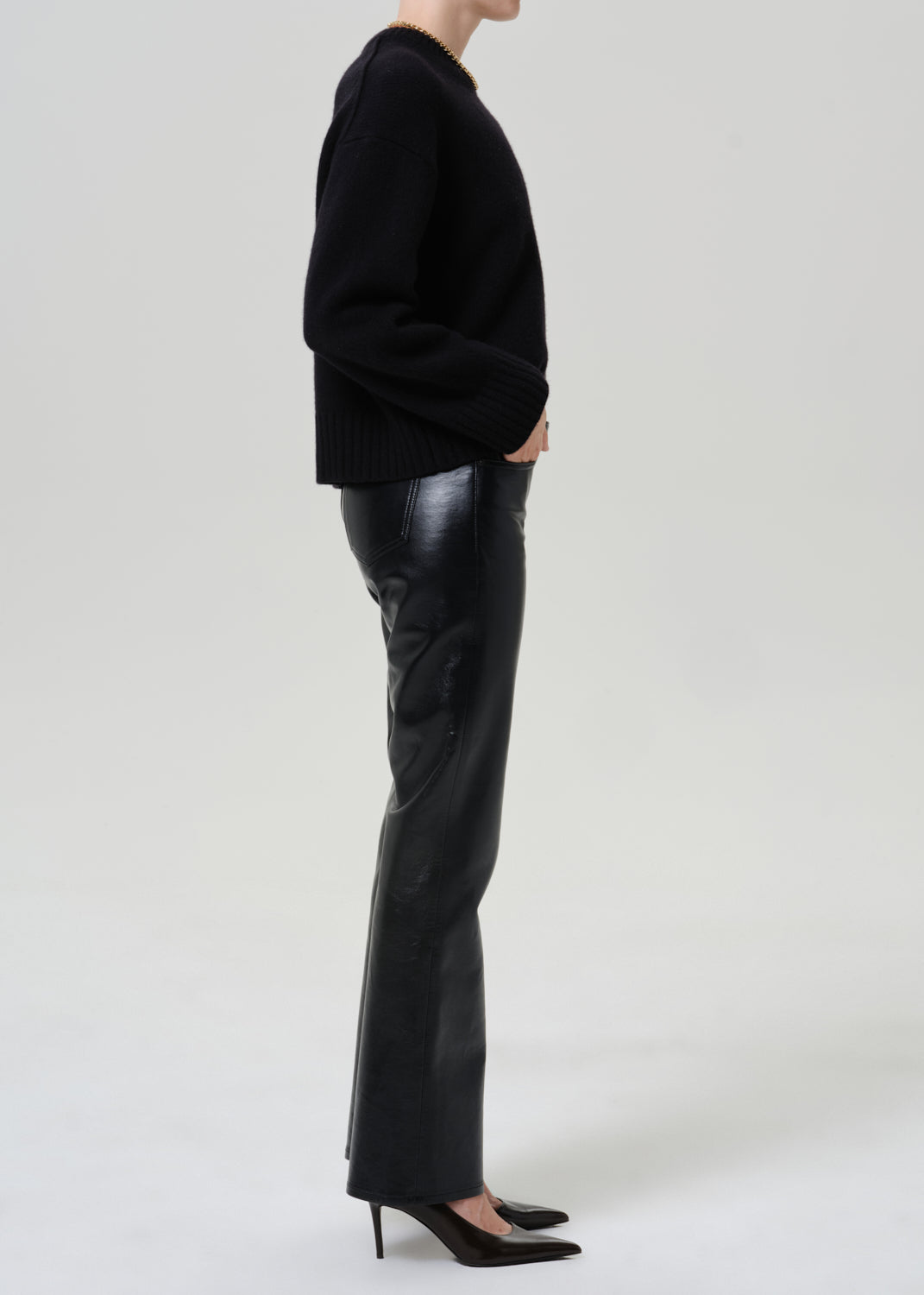 A model shown in profile wearing a black knit sweater, black leather flare pants, and black pointed-toe heels against a light gray studio background.