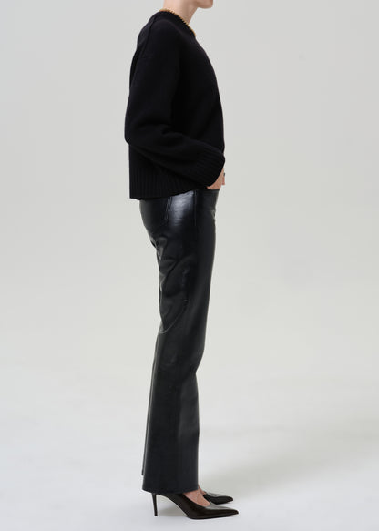 A model shown in profile wearing a black knit sweater, black leather flare pants, and black pointed-toe heels against a light gray studio background.