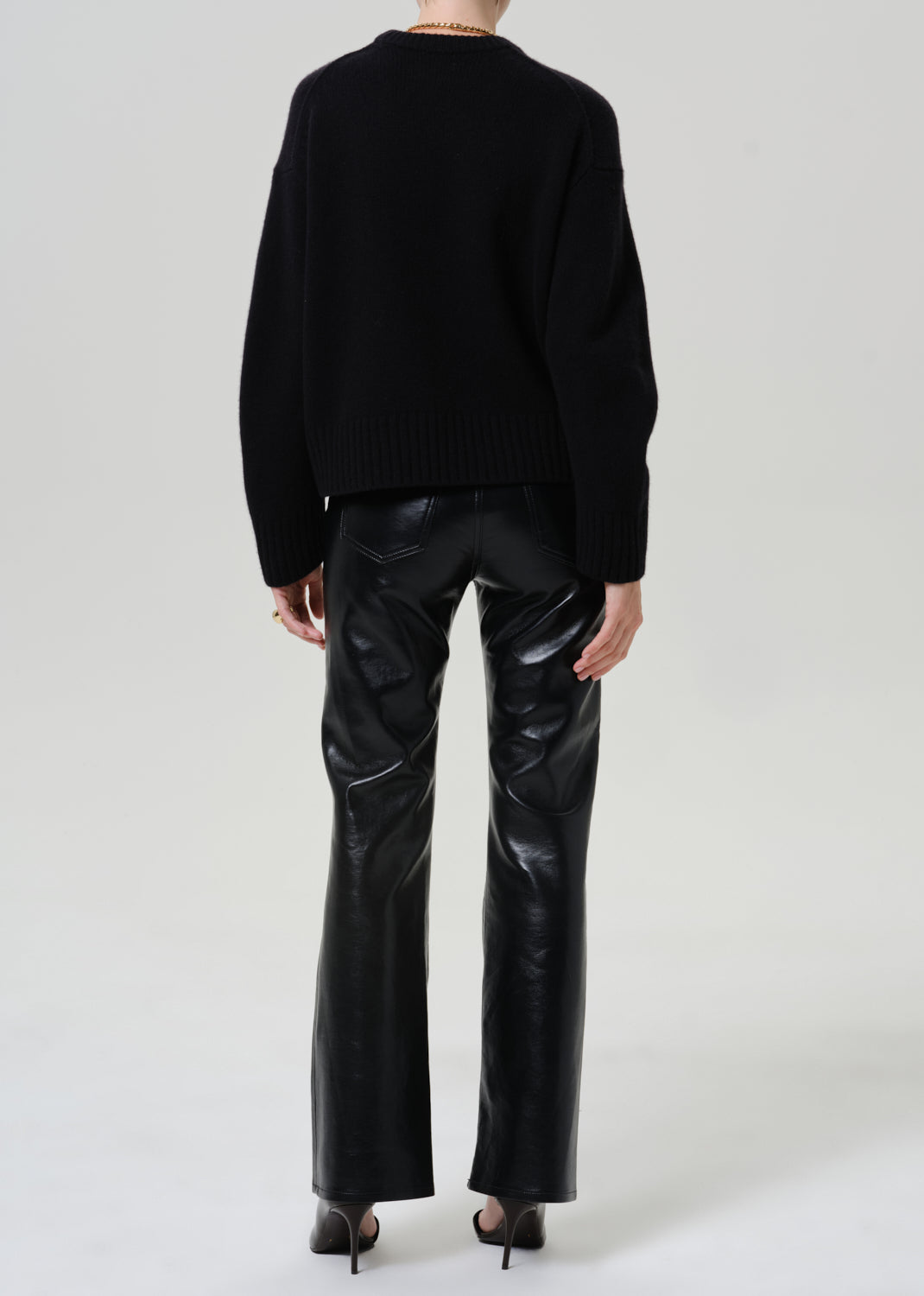 A model shown from the back wearing a black knit sweater and shiny black leather flare pants, paired with black high heels, against a light gray studio background.