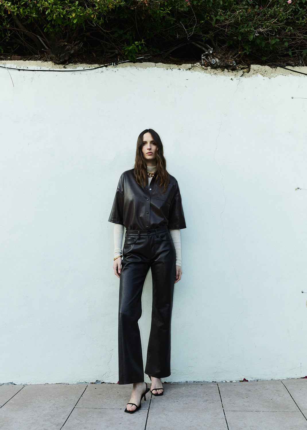 Woman in brown leather outfit standing against a white wall with greenery at the top.