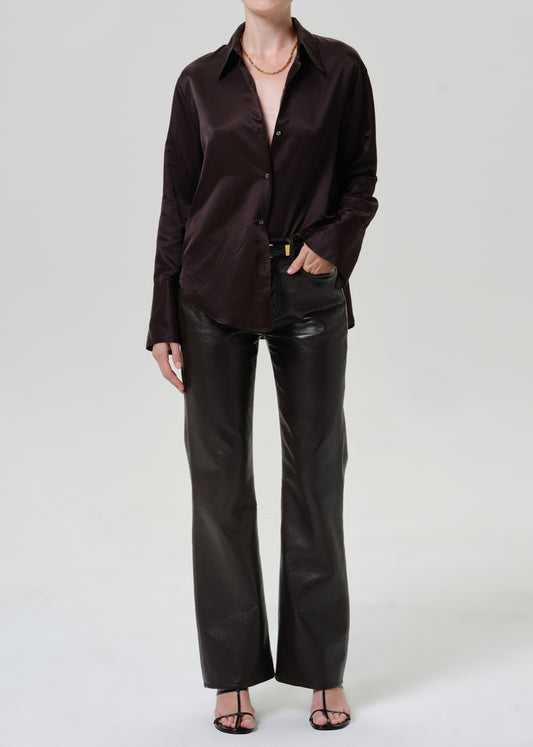 A model wearing a dark brown satin button-down shirt and black faux-leather wide-leg pants, styled with a gold necklace and black sandals against a light gray studio background.