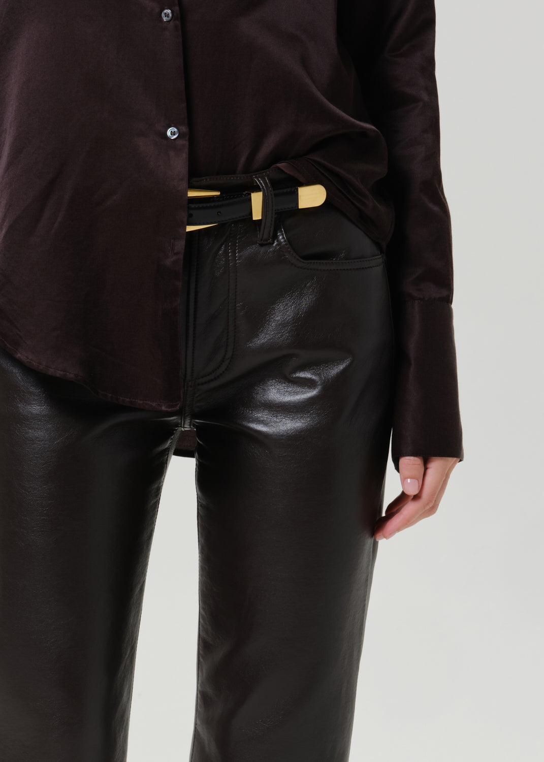 A close-up of a person wearing a dark brown satin button-up shirt and shiny black faux-leather pants, styled with a black belt featuring gold-tone accents.