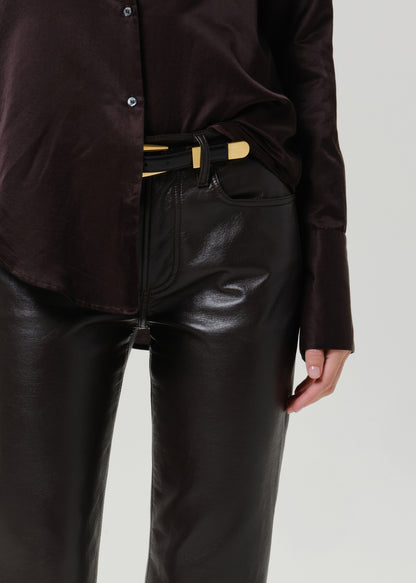 A close-up of a person wearing a dark brown satin button-up shirt and shiny black faux-leather pants, styled with a black belt featuring gold-tone accents.