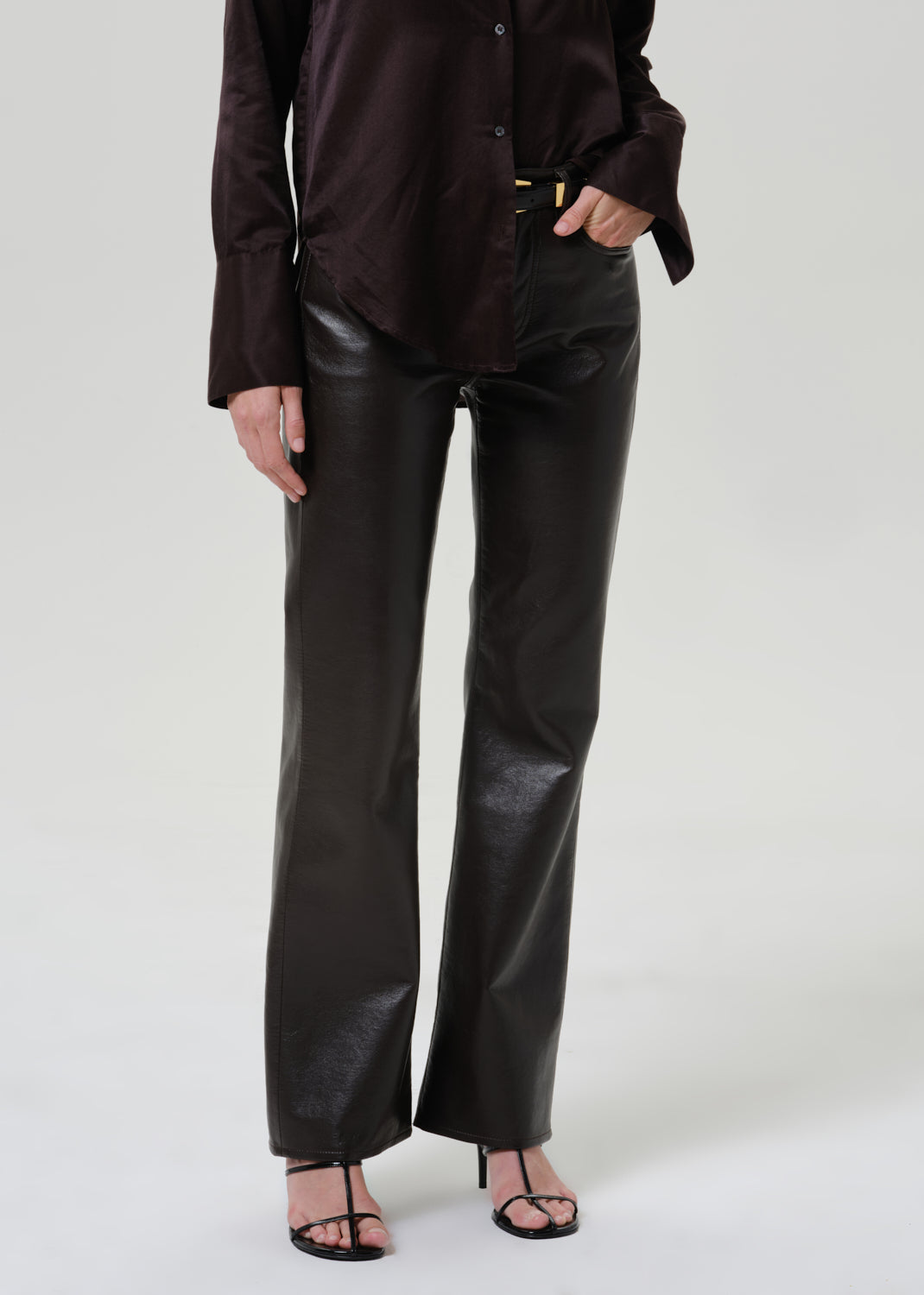 A model wearing black faux-leather wide-leg trousers paired with a dark brown satin blouse and black strappy heels, shown from the waist down against a light gray studio background.
