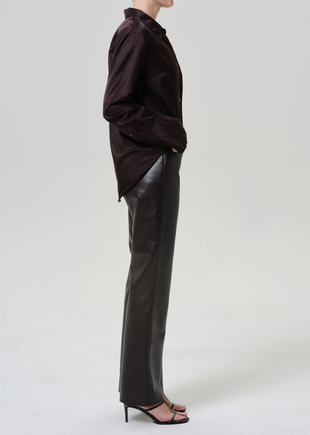 A model shown in profile wearing a dark brown button-up shirt and black leather trousers, paired with black high-heeled sandals, against a light gray studio background.