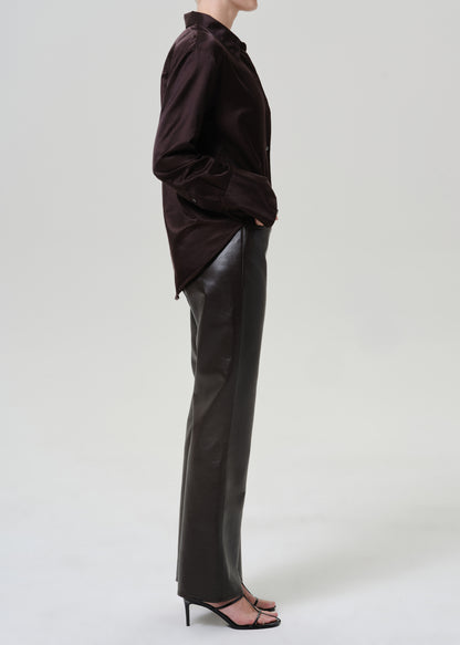 A model shown in profile wearing a dark brown button-up shirt and black leather trousers, paired with black high-heeled sandals, against a light gray studio background.