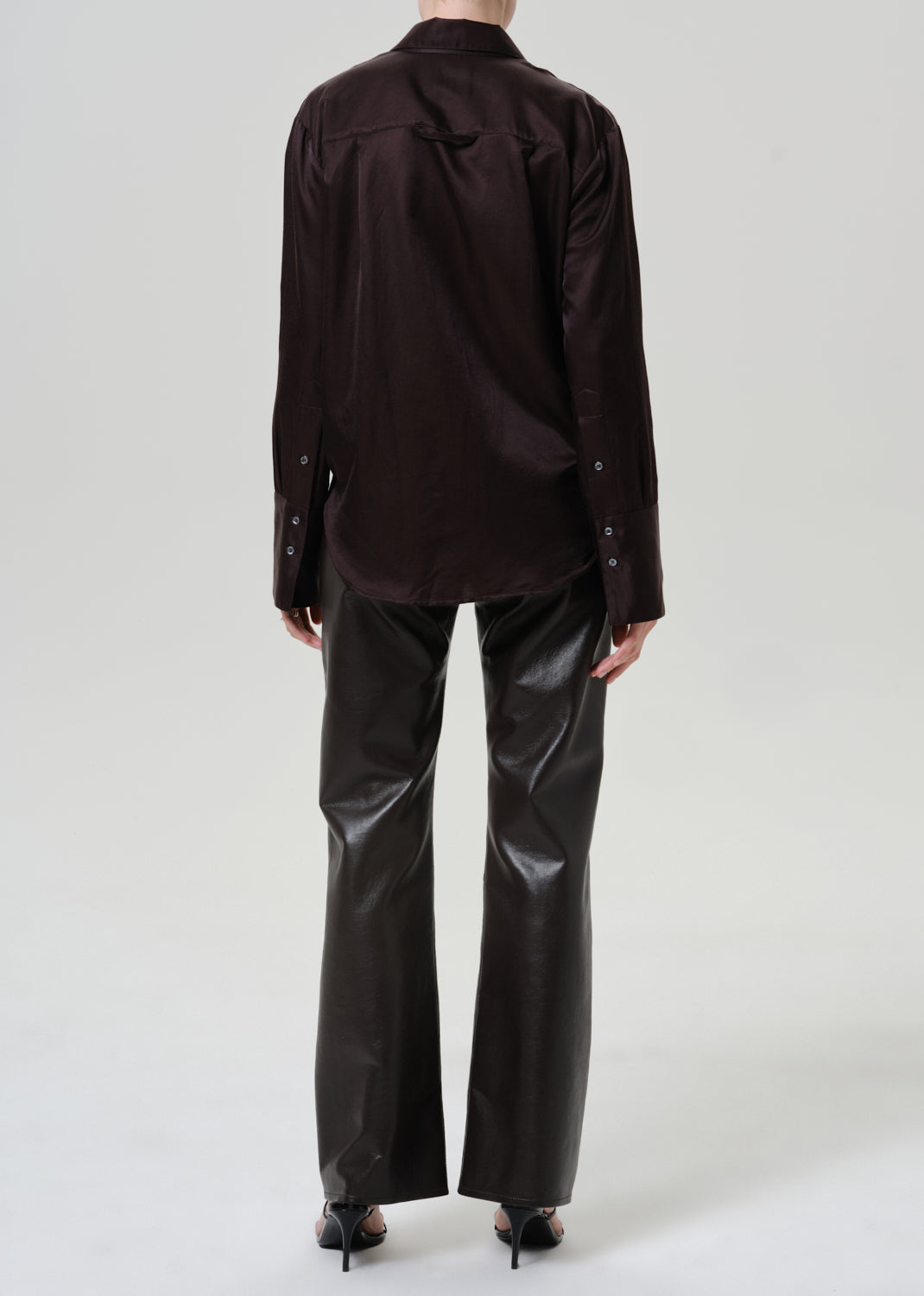 A model shown from the back wearing a dark brown long-sleeve button-down shirt and shiny black wide-leg trousers, paired with black high heels, against a light gray studio background.