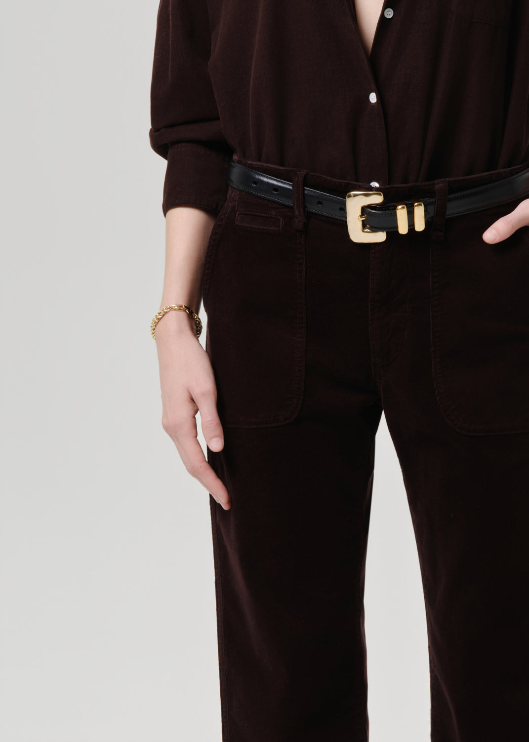 Person wearing a dark brown outfit with a black belt and gold buckle on a grey background