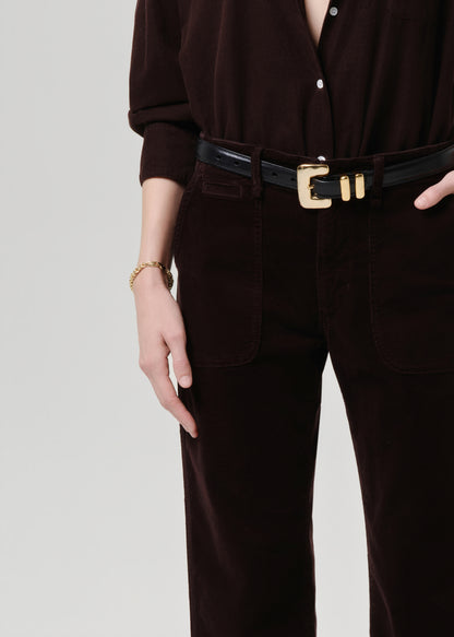 Person wearing a dark brown outfit with a black belt and gold buckle on a grey background