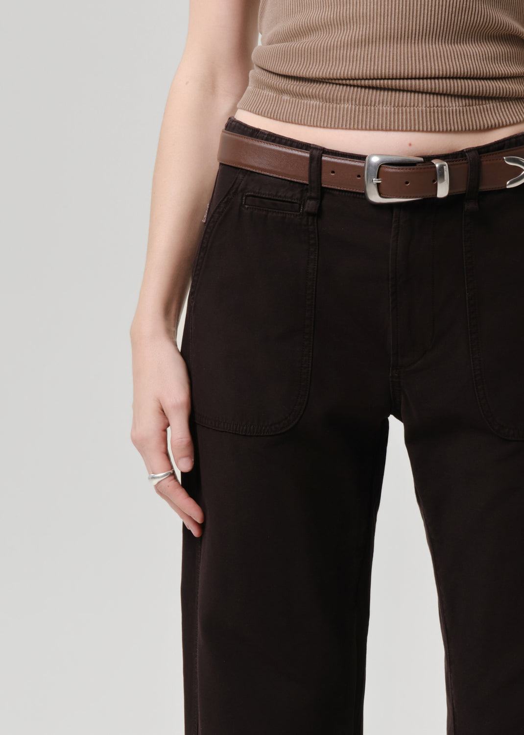 Person wearing brown ribbed top and dark pants with a belt on a grey background