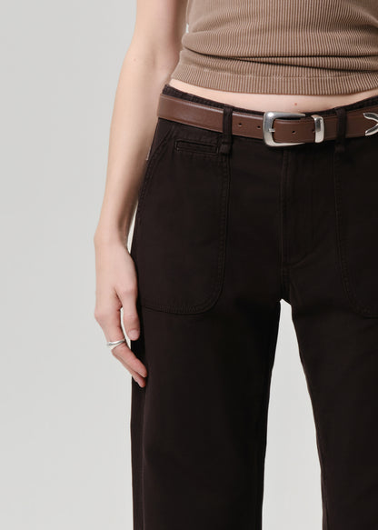 Person wearing brown ribbed top and dark pants with a belt on a grey background