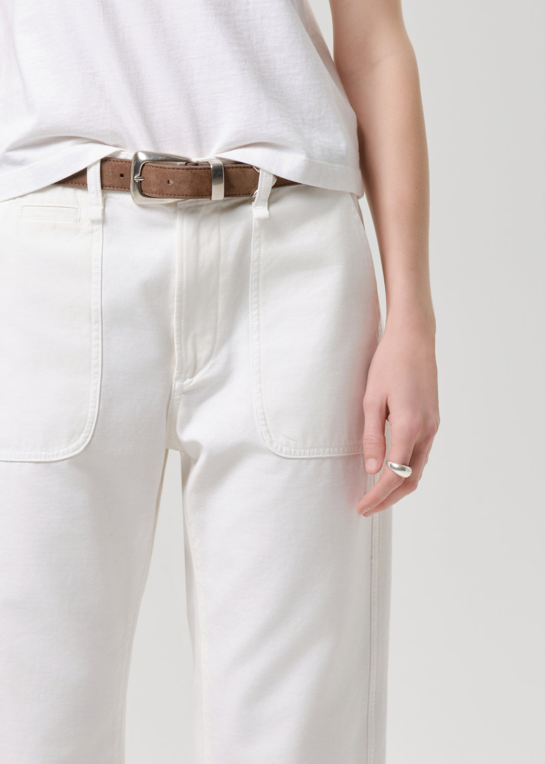 A close-up of a person wearing off-white wide-leg trousers with a brown leather belt and a white sleeveless top, against a grey background.