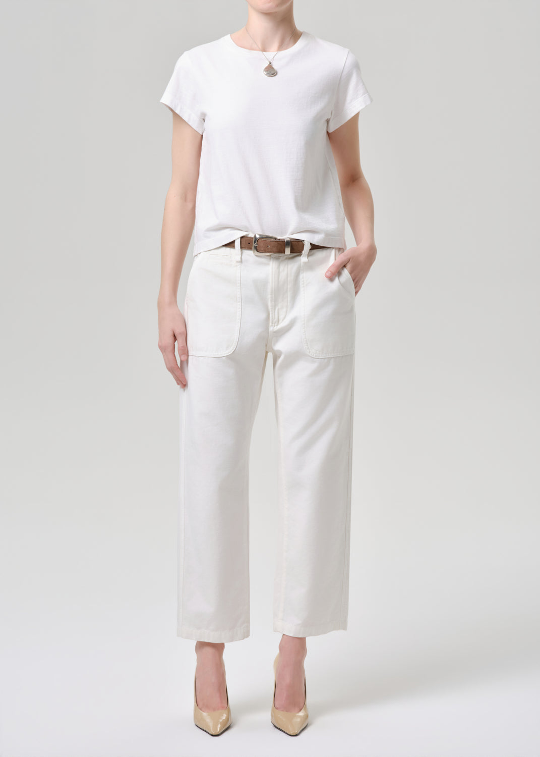 A model wearing a white short-sleeve crewneck t-shirt, white cropped wide-leg pants with front patch pockets, a brown leather belt, and beige pointed-toe heels, standing against a grey background.