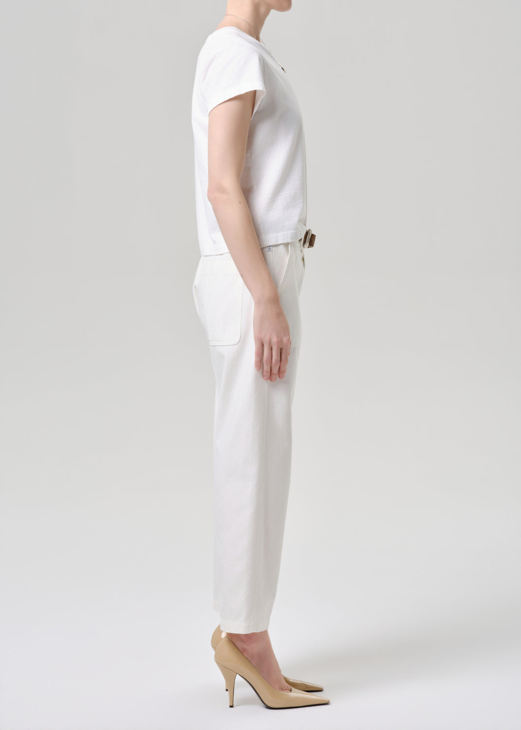 A model shown in profile wearing a white short-sleeve top, white trousers, and beige high heels against a grey background.