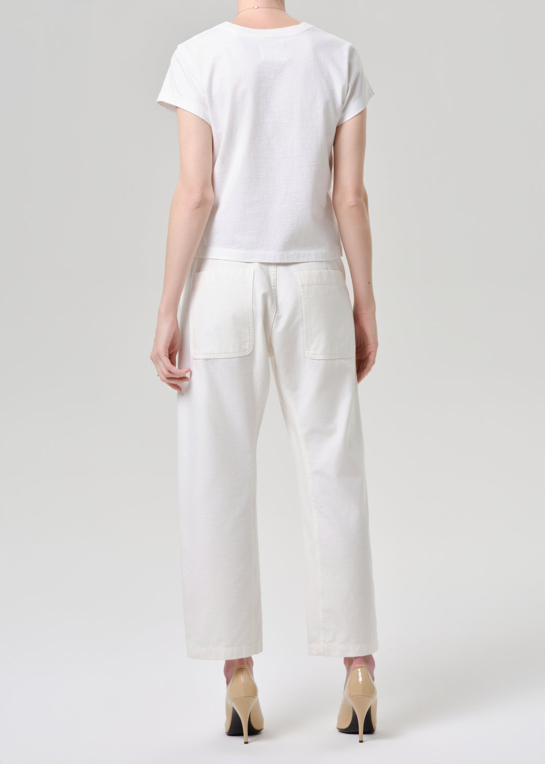 A model shown from the back wearing a white short-sleeve t-shirt and white wide-leg cropped pants, paired with beige high heels, against a grey background.