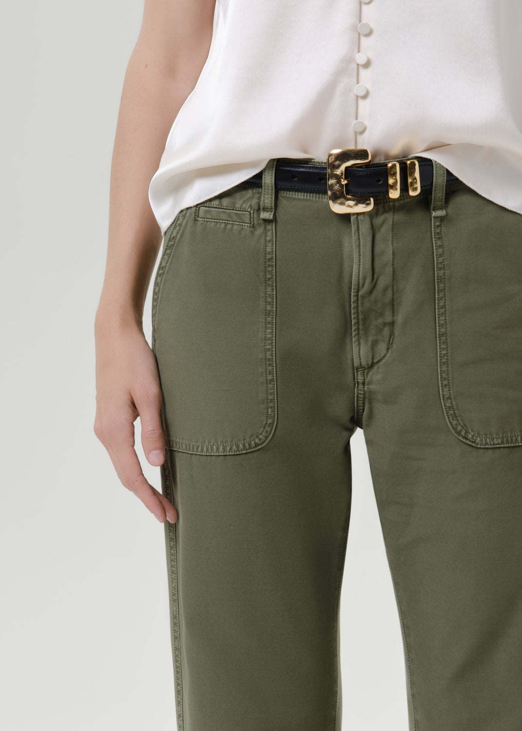 Close-up of a person wearing olive green pants with a black belt featuring a prominent gold buckle, paired with a white top against a grey background.