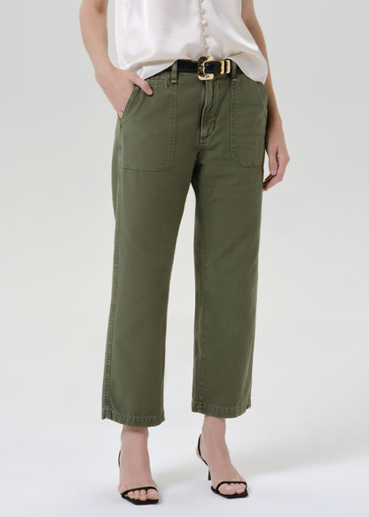A model wearing olive green cropped pants with a black belt featuring a gold buckle, styled with a white blouse and black heeled sandals against a grey background.