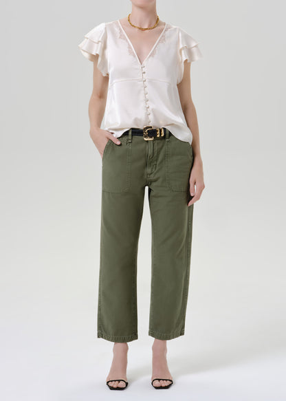 A model wearing a white ruffled short-sleeve blouse and olive green cropped pants with a black belt, standing against a grey background.