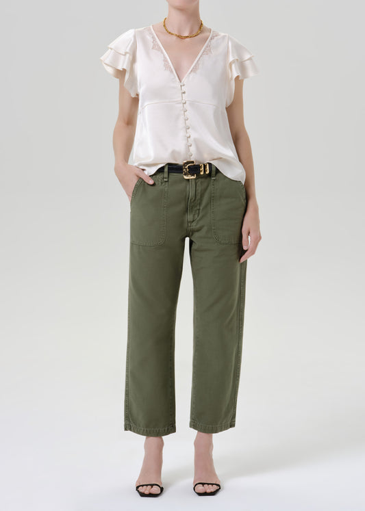 A model wearing a white ruffled short-sleeve blouse and olive green cropped pants with a black belt, standing against a grey background.