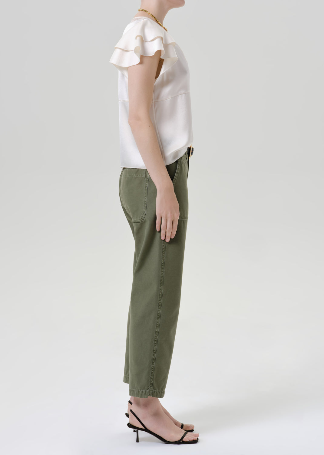 A model shown in profile wearing olive green cropped trousers, a white ruffled short-sleeve blouse, and black strappy high-heeled sandals against a grey background.