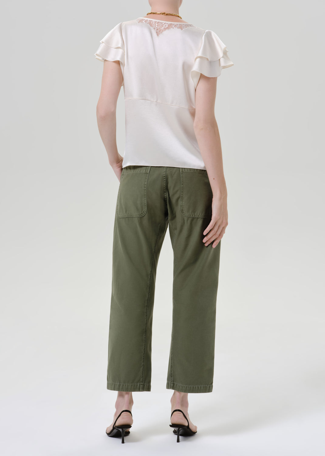 A model shown from the back wearing olive green cropped pants, a cream ruffled short-sleeve blouse, and black heeled sandals against a grey background.