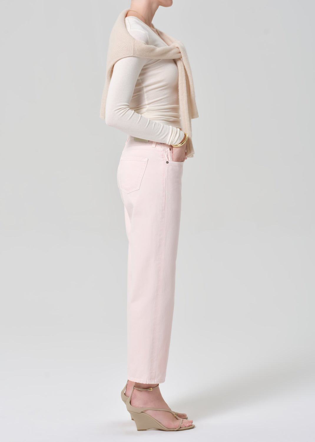 Person wearing a cream top, beige sweater, and light pink jeans on a grey background