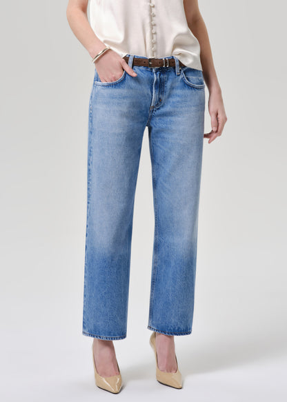 A model wearing light blue cropped straight-leg jeans with a brown belt, paired with a cream blouse and beige heels, against a grey background.