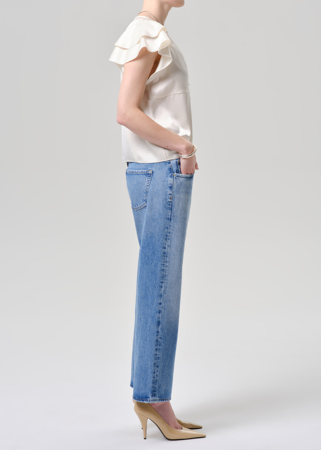 A model shown in profile wearing a cream ruffled short-sleeve top, light-wash blue straight-leg jeans, and beige high heels against a grey background.