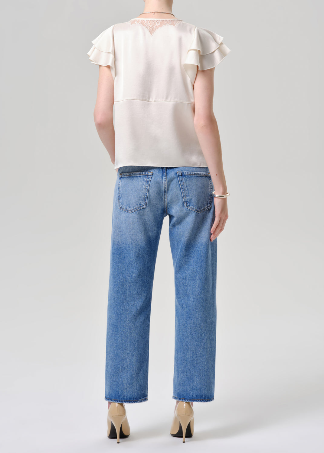 A model shown from the back wearing a cream satin ruffle-sleeve blouse and light blue cropped jeans, paired with beige high heels, against a grey background.