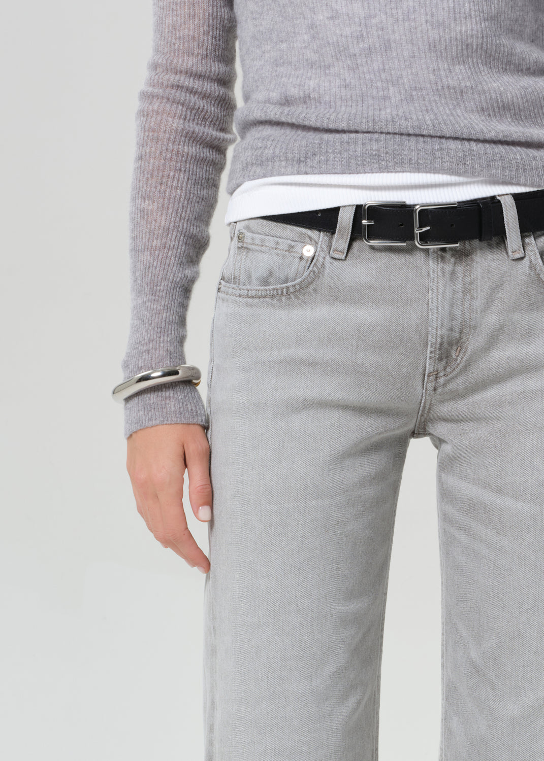 Person wearing a gray sweater, white shirt, black belt, and light gray jeans on a plain background