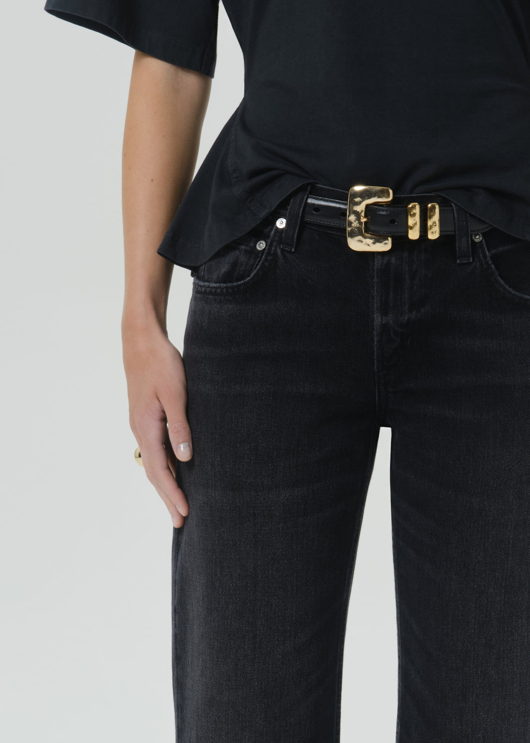 Close-up of a person wearing a black belt with a large gold-tone buckle over dark washed jeans and a black top, against a grey background.