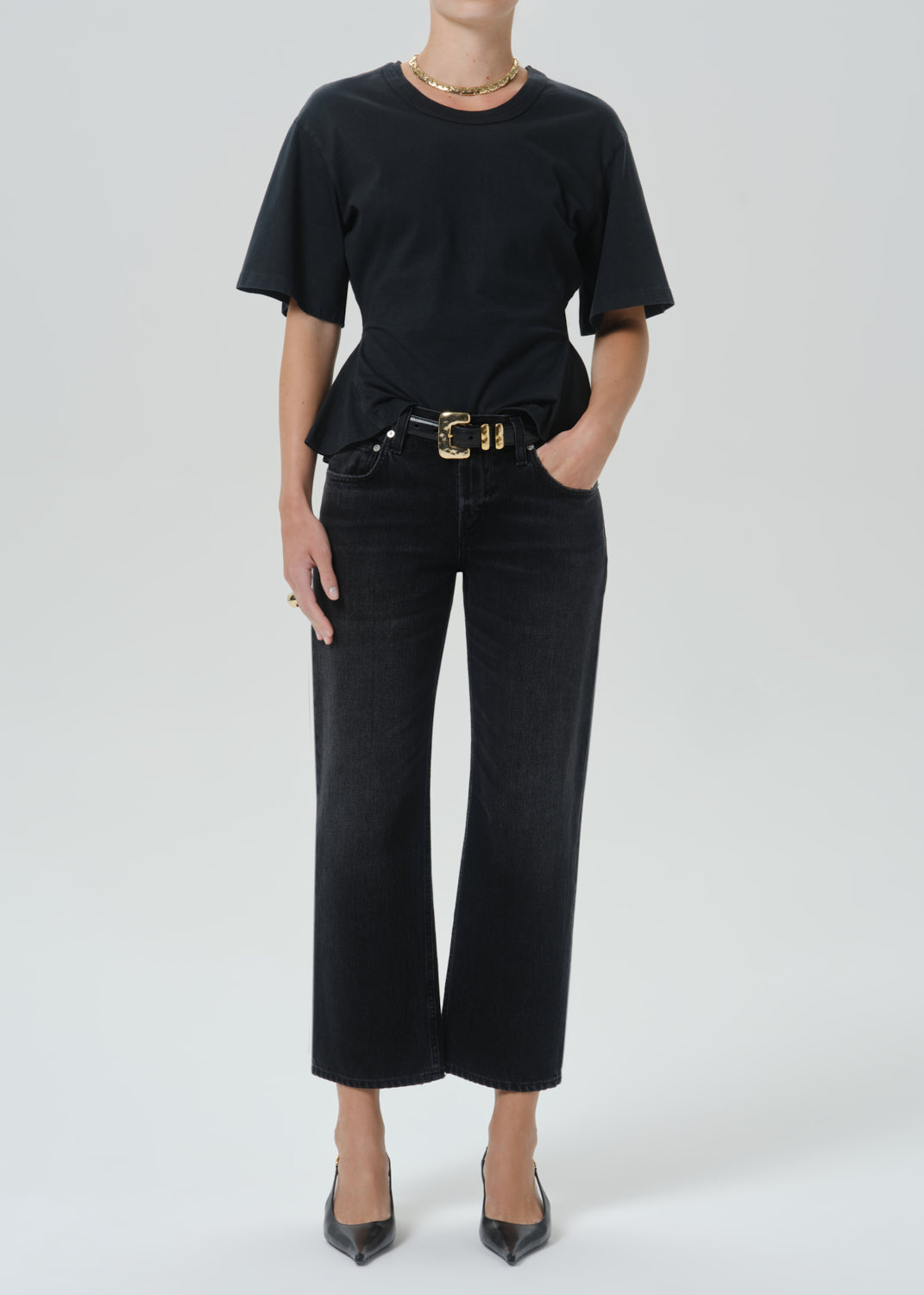 A model wearing a black short-sleeve crewneck t-shirt, black cropped wide-leg jeans with a gold-tone belt, and black pointed-toe heels against a grey background.