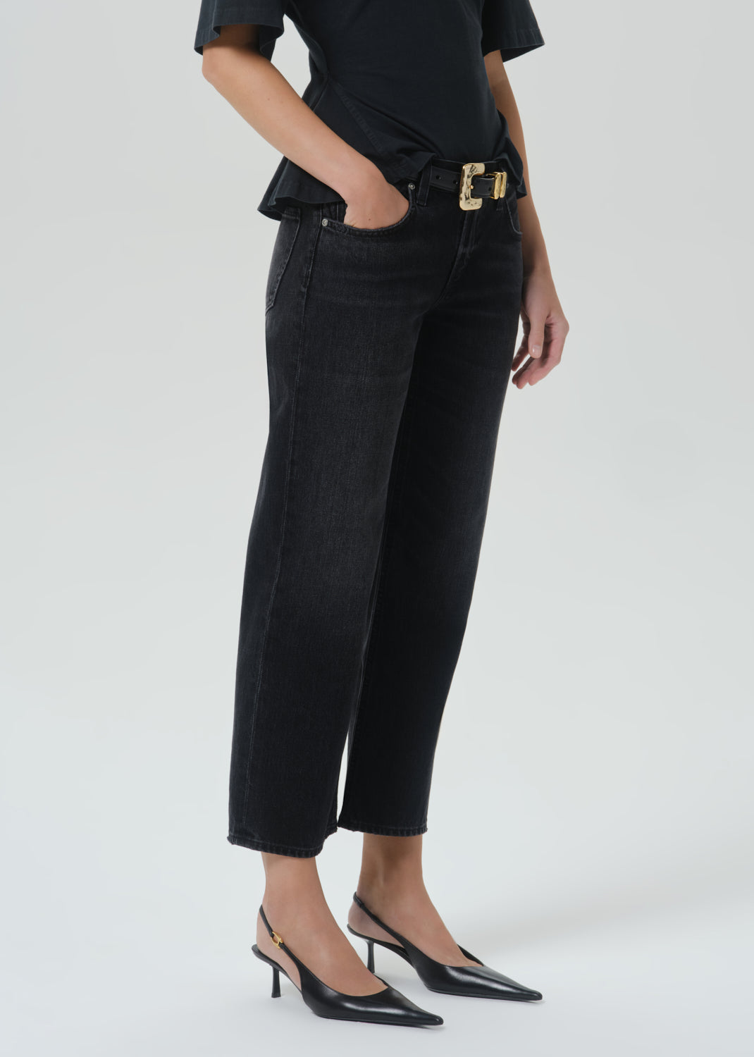 A model wearing black cropped jeans with a gold-tone belt buckle, paired with a black top and black slingback heels, shown from the waist down on a grey background.