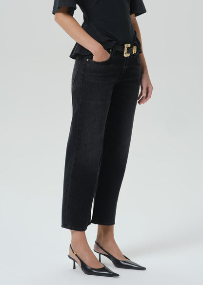 A model wearing black cropped jeans with a gold-tone belt buckle, paired with a black top and black slingback heels, shown from the waist down on a grey background.