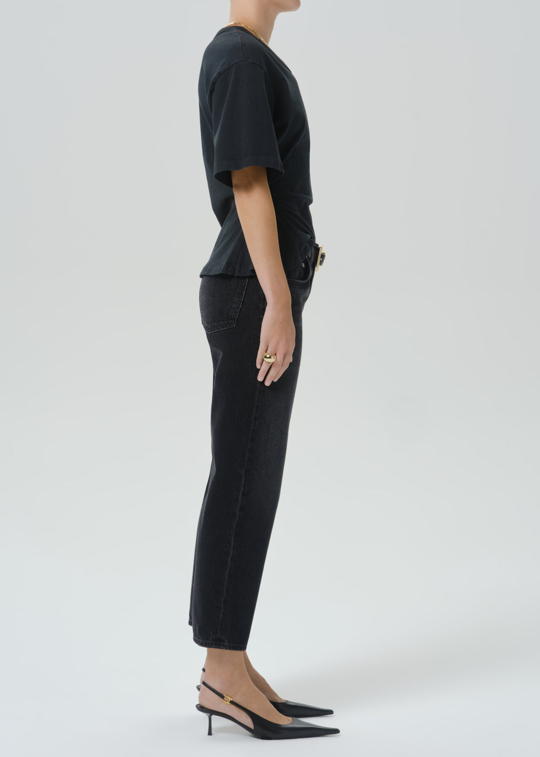 A model shown in profile wearing a black short-sleeve top, black cropped pants, and black slingback heels against a grey background.