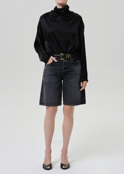 Woman wearing a black satin draped neck top with black denim shorts on a grey background.