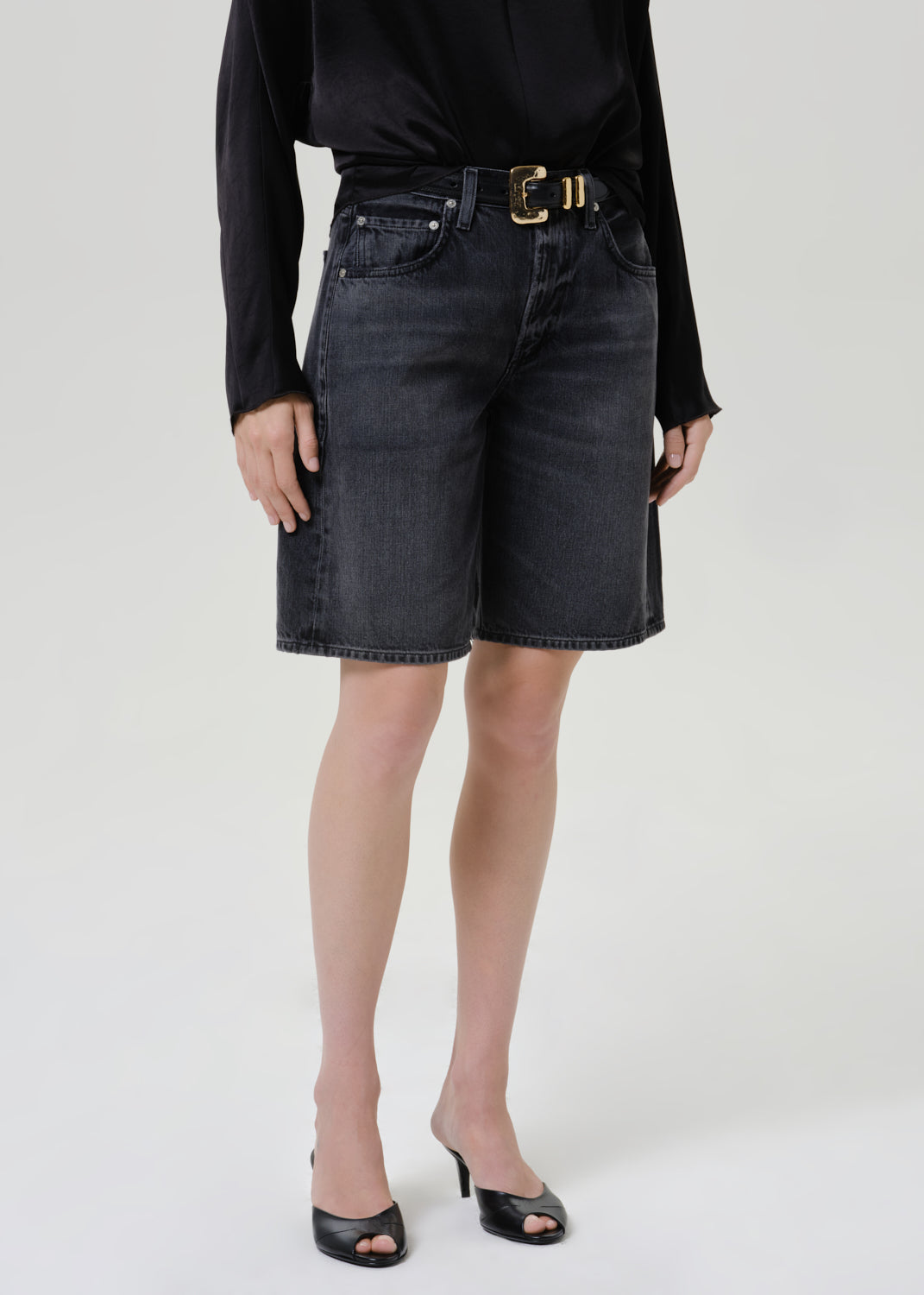 Woman wearing a black satin draped neck top with black denim shorts on a grey background.