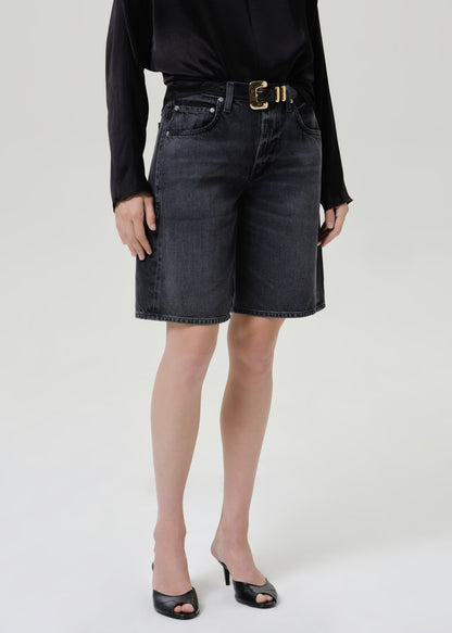 Woman wearing a black satin draped neck top with black denim shorts on a grey background.
