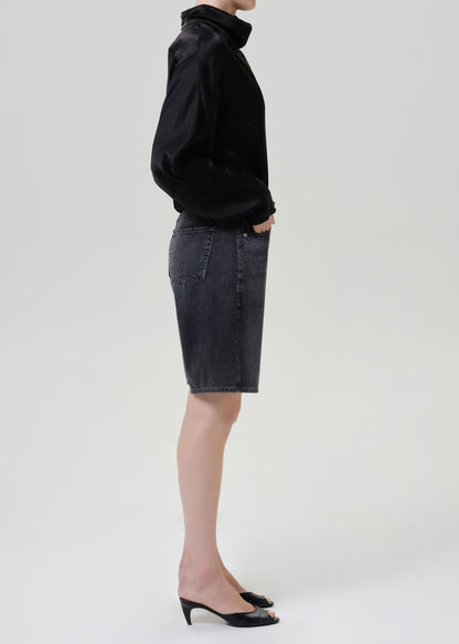 Woman wearing a black satin draped neck top with black denim shorts viewed from the side on a grey background.