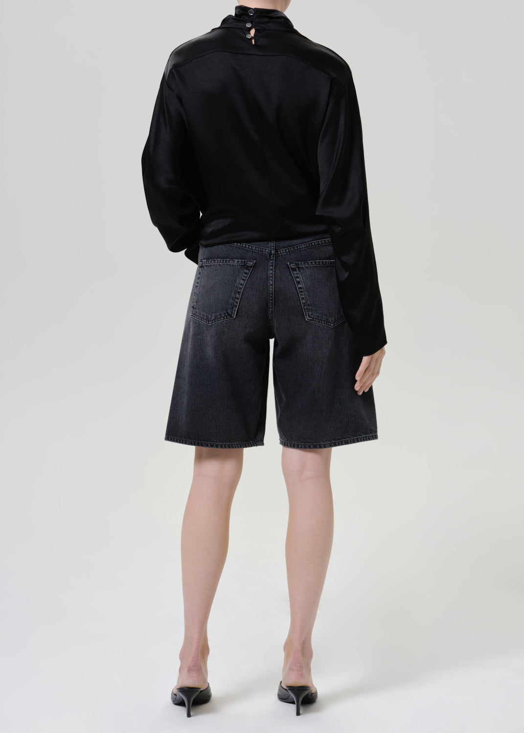 Woman wearing a black satin draped neck top with black denim shorts viewed from the back on a grey background.