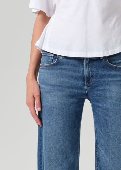 Close-up of a person wearing medium-wash blue jeans and a white short-sleeve top against a grey background.